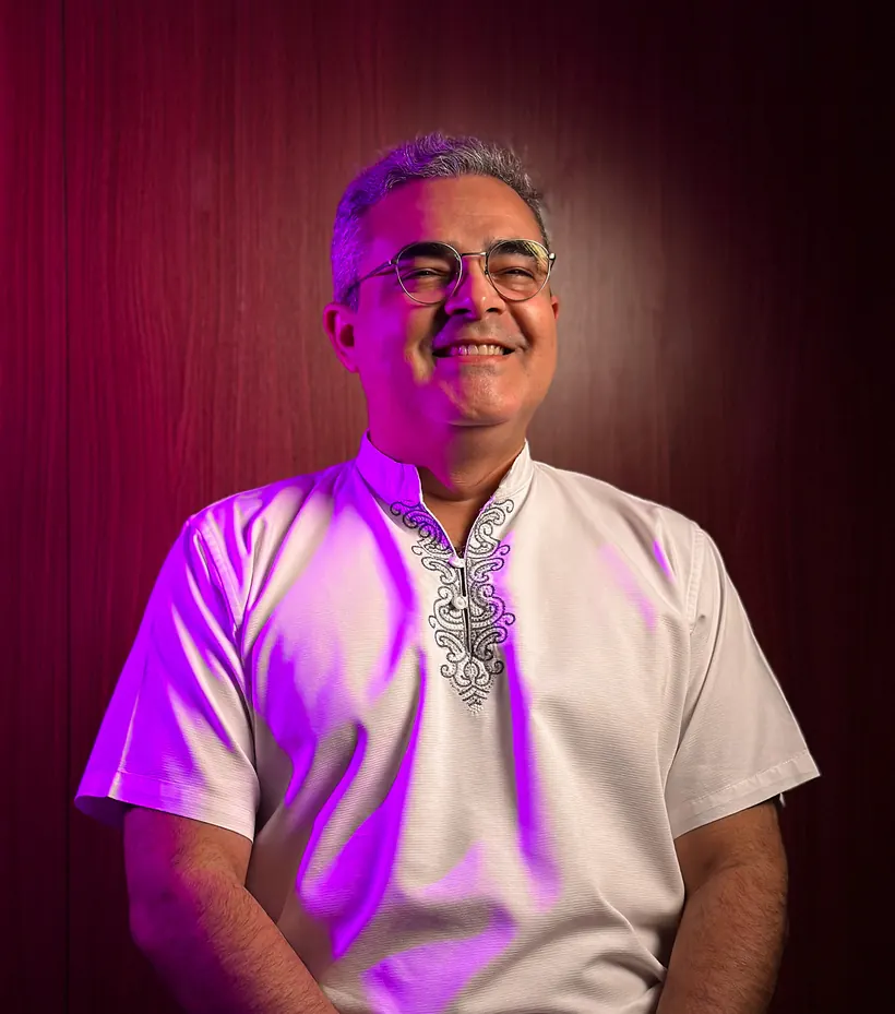 Sidnei Bonfim rindo, sentado, de camisa branca bordada, sob uma luz magenta